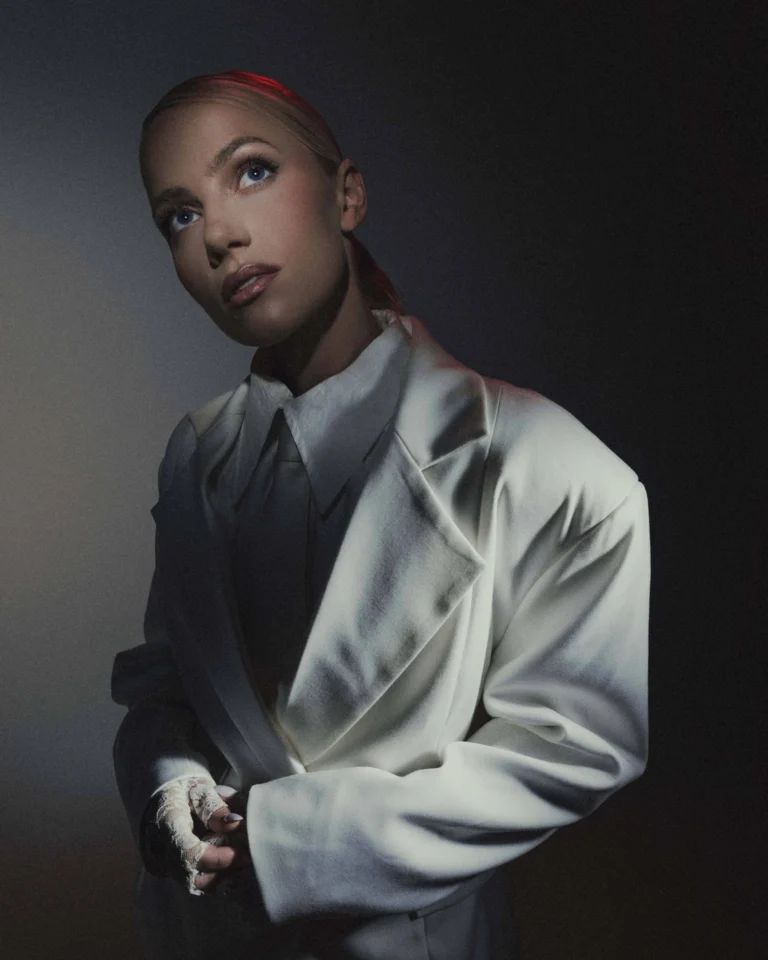Woman with blonde hair wearing an oversized white suit and lace glove, gazing upwards.
