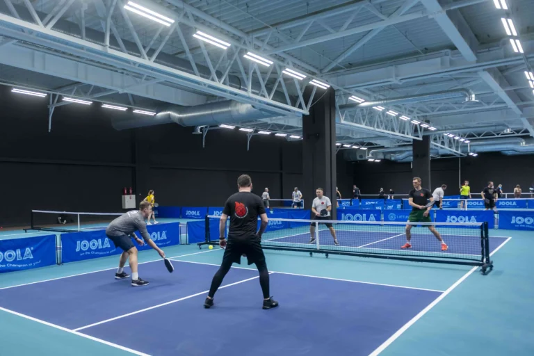 Men playing pickleball on a blue court inside a large sports facility with multiple matches.