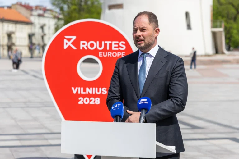 A man in a suit speaks at a podium with microphones before a large red sign.