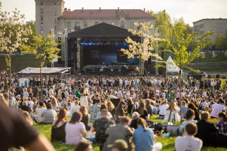 A large crowd sits on a grassy hill facing an outdoor concert stage during the day.