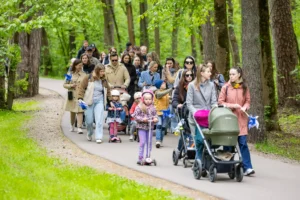 Families with strollers and kids on scooters walk on a paved path through a green forest.