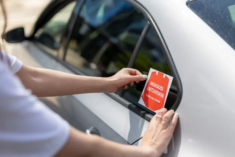 Hands applying a red 'learning Lithuanian' sticker to the rear window of a silver car.
