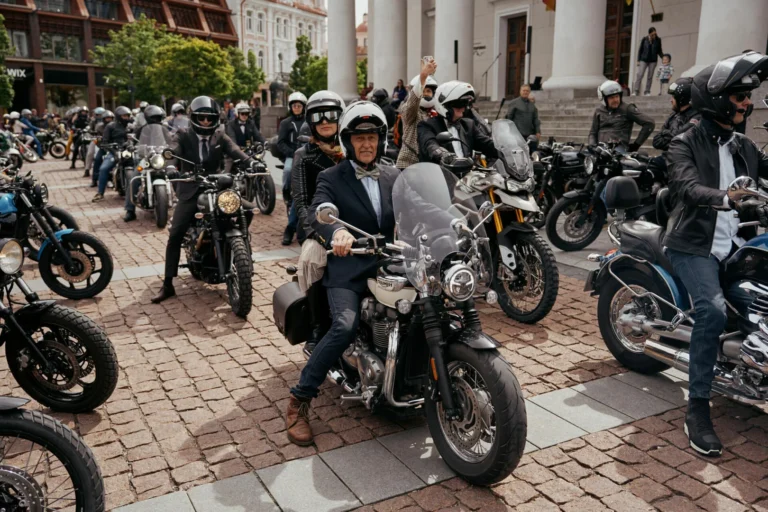 Motorcyclists in formal suits and helmets gather on a cobblestone square with vintage style motorcycles.