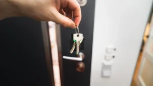 A hand holding a set of keys with a house-shaped keychain in front of a modern apartment door, symbolizing new rental protections for tenants.