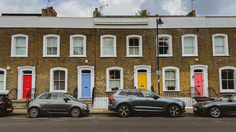A row of London terraced houses with white window frames and colorful doors under a cloudy sky.