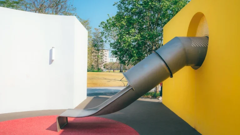 A sleek modern playground slide emerging from a bright yellow wall in a newly designed park area.