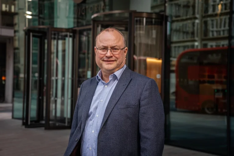 Middle-aged man wearing glasses and a blue blazer standing before a glass building with revolving doors.