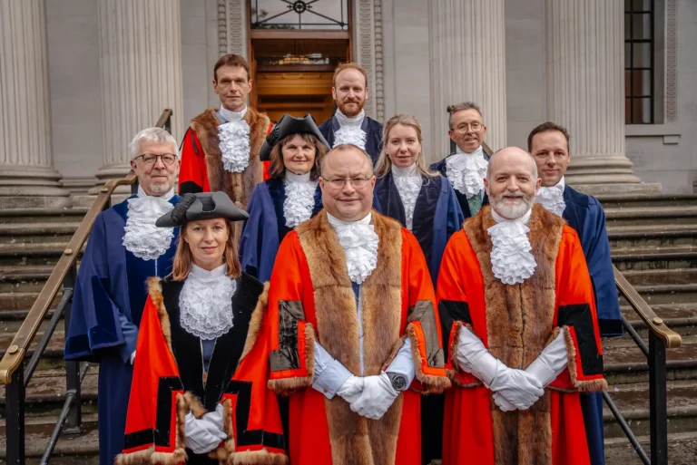 Group of officials in red and blue ceremonial robes with lace ruffs on stone steps.