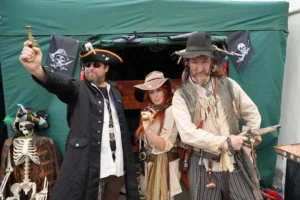 Three people dressed in detailed pirate costumes pose in front of a green tent with pirate flags.