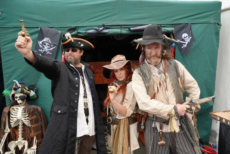 Three people dressed in detailed pirate costumes pose in front of a green tent with pirate flags.