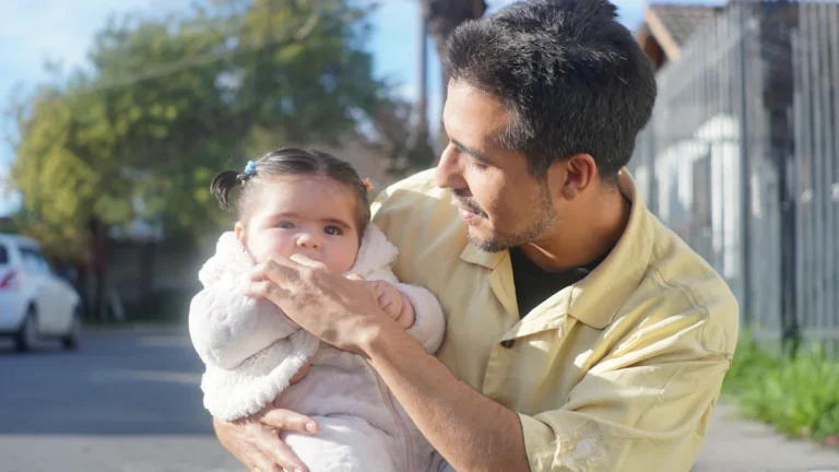 A father holds his young child in his arms outdoors during a bright, sunny day.