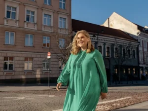 Smiling woman in a long-sleeved green dress walking through a sunlit European city square.