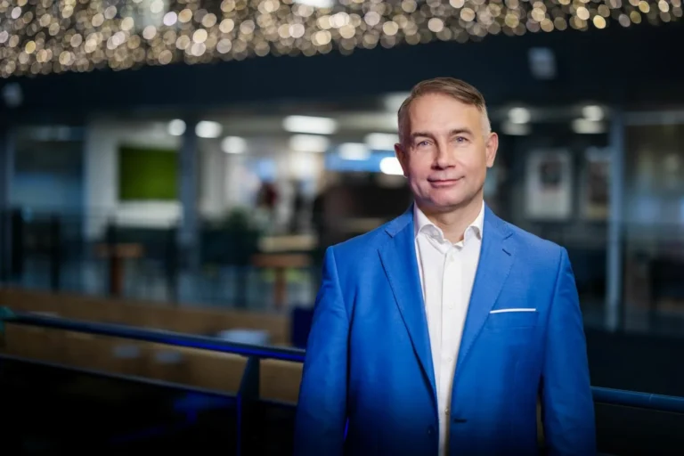 A smiling man wearing a bright blue blazer and white shirt in a modern indoor setting.