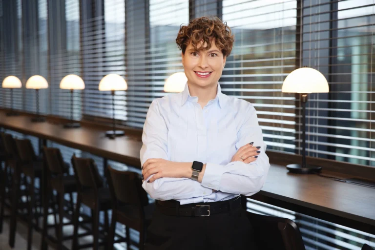 Smiling woman with curly hair stands with arms crossed in a modern office with row lamps.