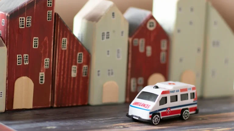 A miniature toy ambulance parked in front of several small wooden toy houses on a wooden surface.