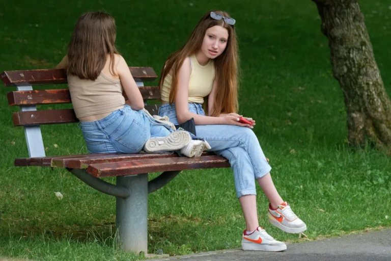 Two teenage girls in jeans sitting on a wooden park bench with green grass behind them.