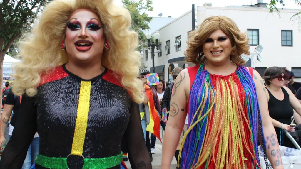 Two drag performers in colorful sequined outfits and wigs walking during a vibrant outdoor Pride parade.