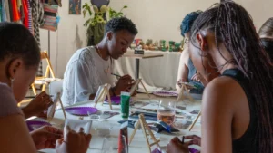 A diverse group of people sitting together at a long table during a community painting workshop, focused on their individual canvases in a social setting.