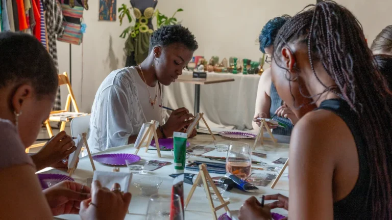 A diverse group of people sitting together at a long table during a community painting workshop, focused on their individual canvases in a social setting.
