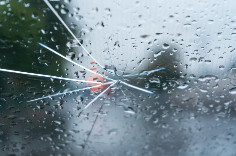 A cracked glass windshield covered in raindrops with blurred orange lights in the background.