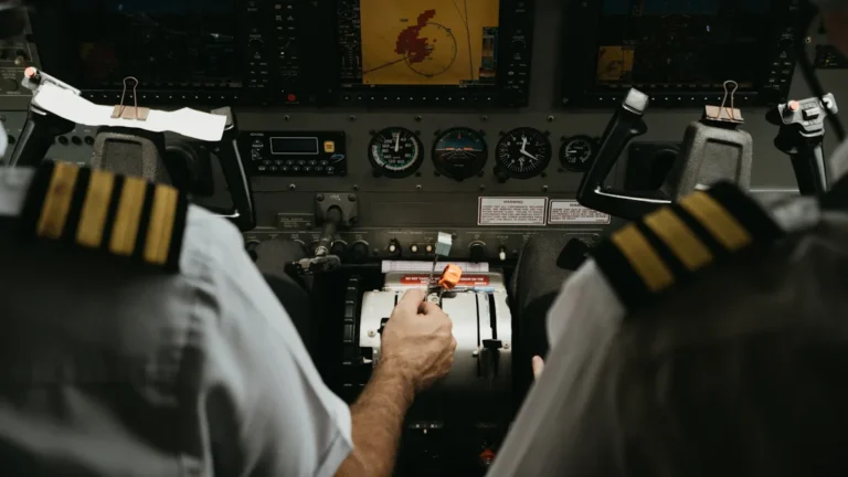 Two commercial pilots in uniform sit in an airplane cockpit with hands on flight controls.