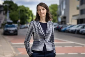 Woman in a striped blazer stands on a street with blurred cars in the background.