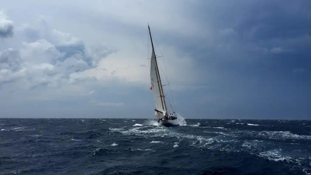 A white sailboat navigating through dark, stormy waters under a cloudy sky.