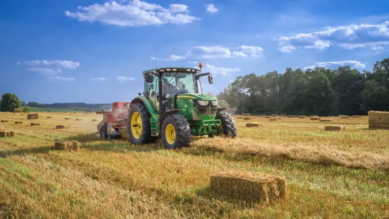 A green tractor working on a farm field with hay bales.