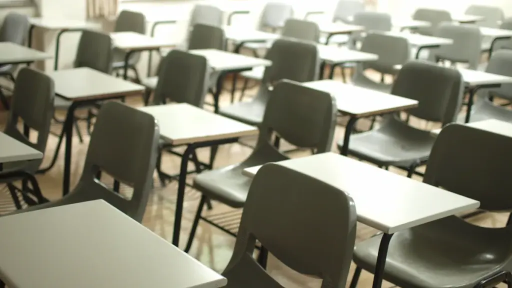 Rows of empty school desks and chairs in a quiet, neutral classroom setting.