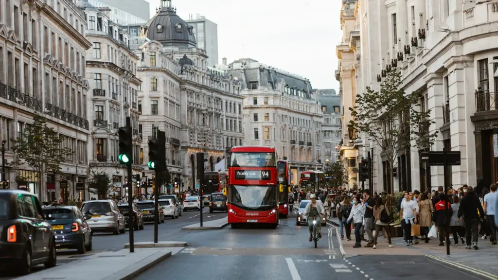 A red double-decker bus driving down a busy London street in summer.