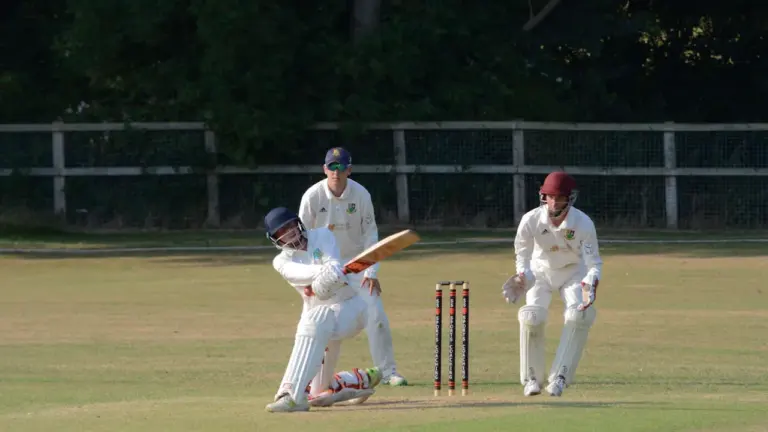Cricket batsman swings bat to hit ball during a competitive match on grass.