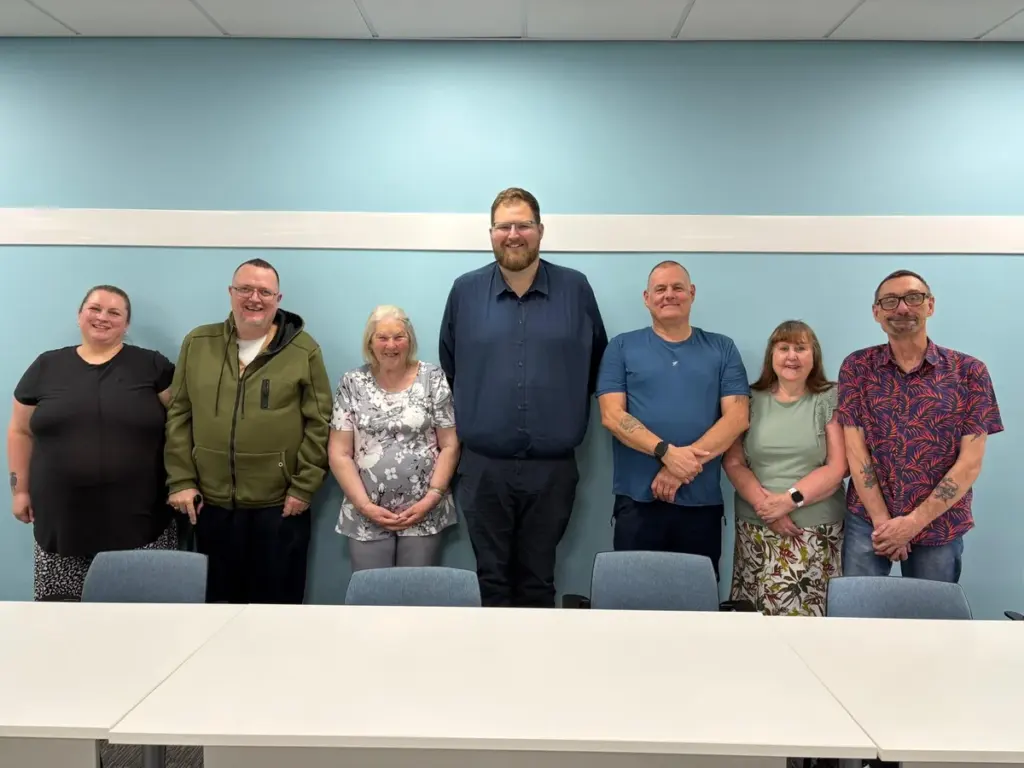 A group of six diverse adults standing in a row against a light blue wall.