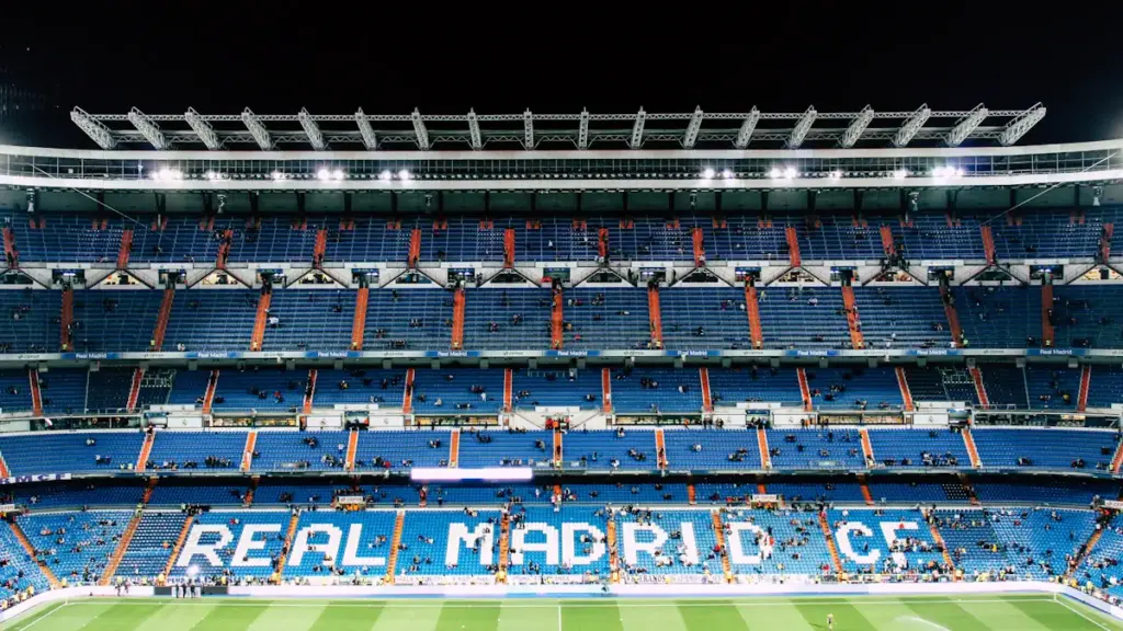 Empty Real Madrid stadium with blue seats and green pitch under bright lights.