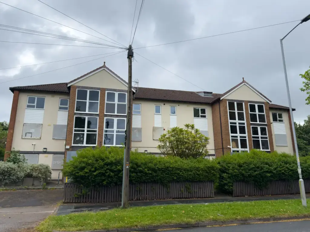 A two-story residential building with boarded-up ground floor windows and overgrown shrubbery outside.
