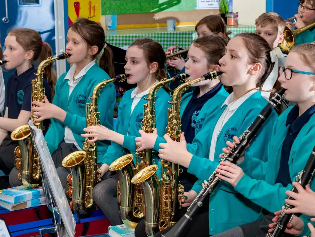 A group of school children in teal cardigans playing saxophones and clarinets in orchestra.