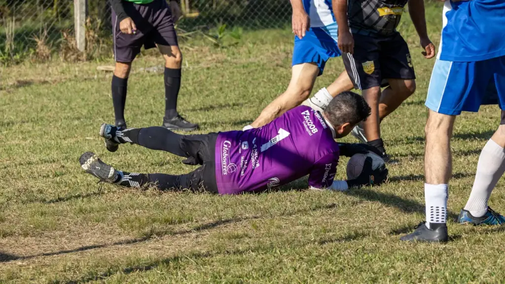A goalkeeper in a purple jersey diving for a soccer ball on grass.