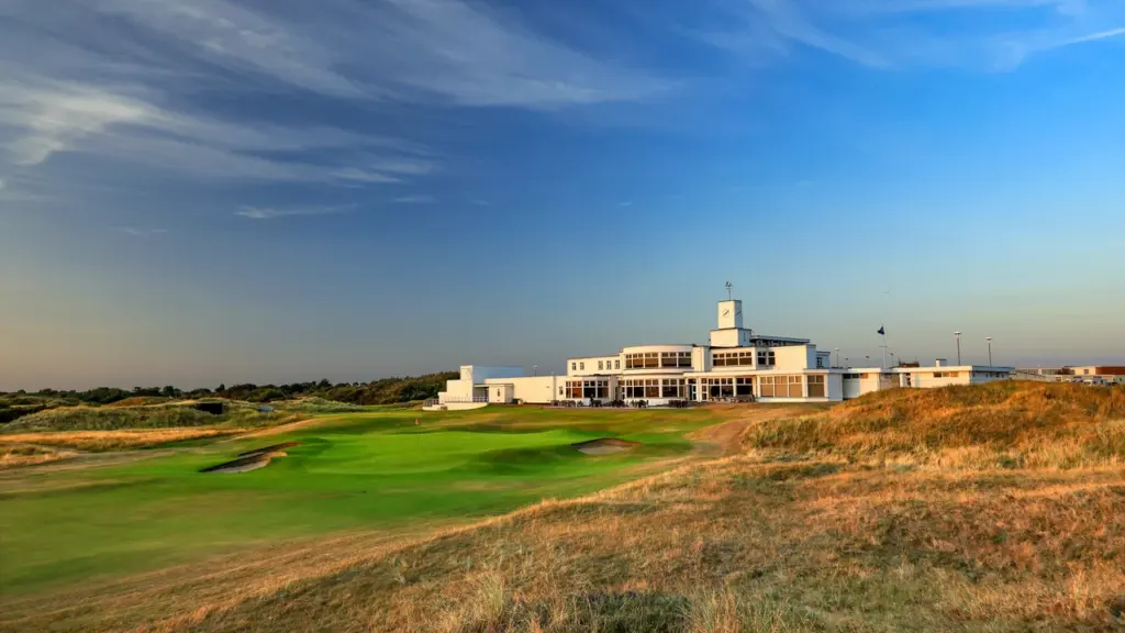 A white golf clubhouse stands behind a green course with sand bunkers under blue skies.