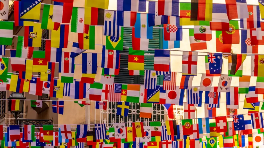 Colorful international flags hanging outdoors against a neutral architectural building facade.
