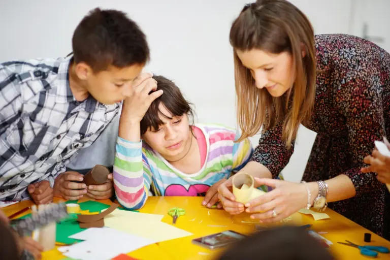 A teacher helps young students with a craft project using paper and cardboard tubes.